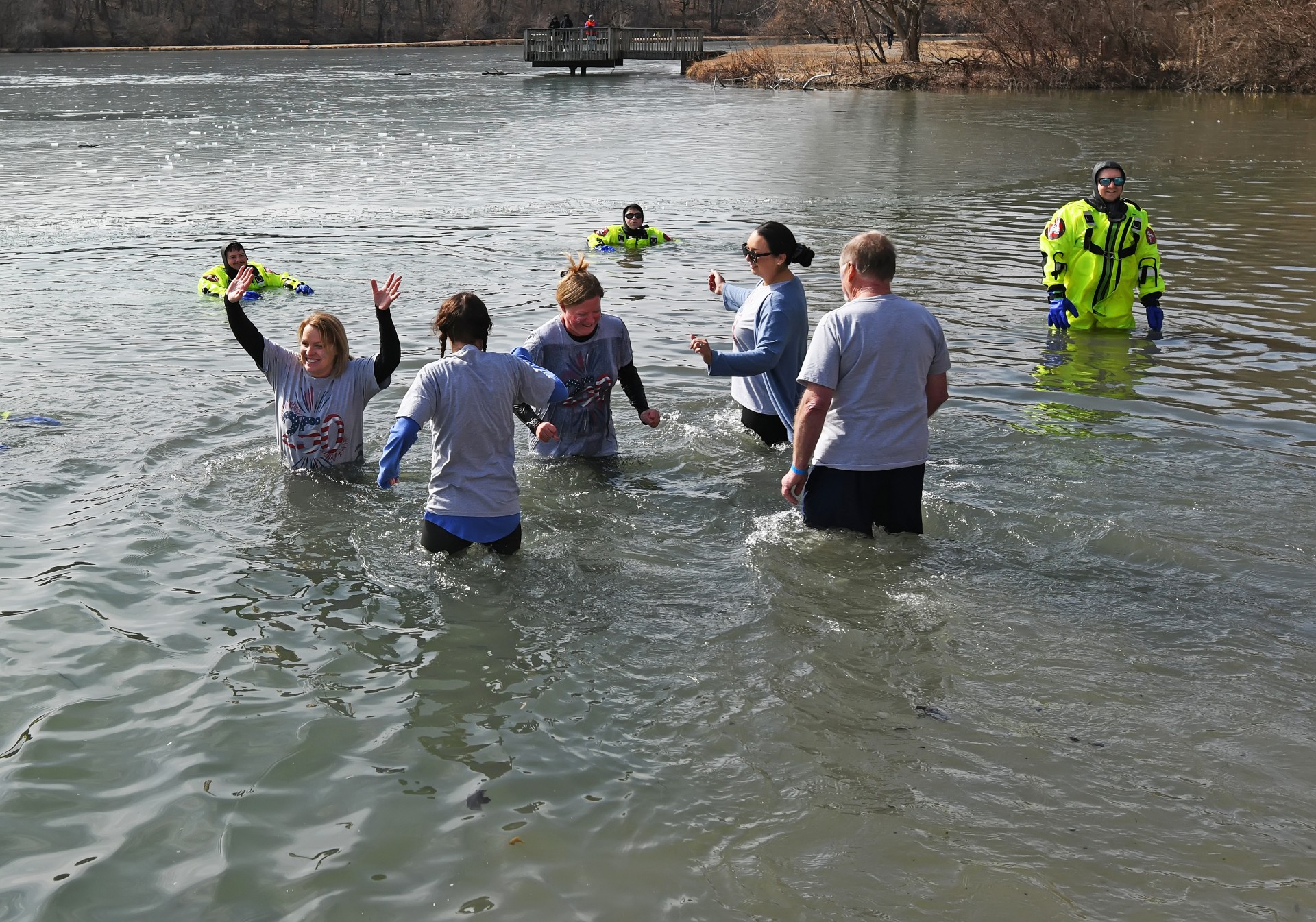 Several people are wading in a cold body of water, with some wearing bright yellow survival suits. A person in a lime green jacket and blue gloves stands on the right side of the frame, appearing to be an attendant.
