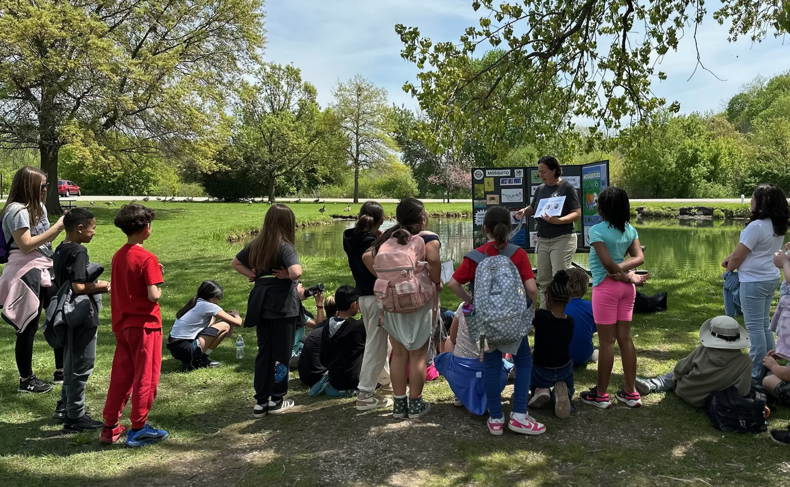 A group of children gathered around an adult presenter in front of a pond, listening to an outdoor lesson with educational posters displayed.