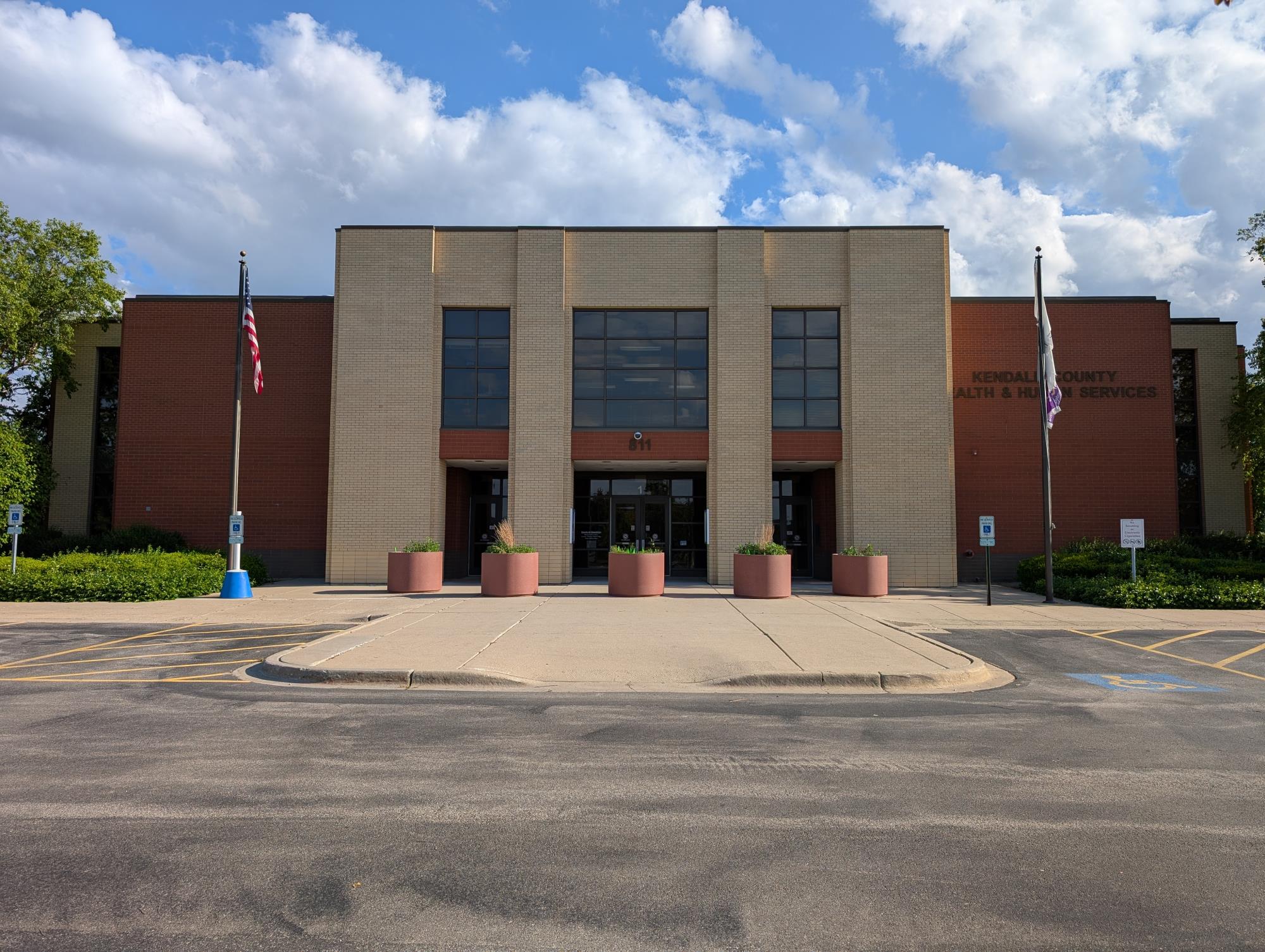 The front entrance of the Kendall County Health Department building features a brick-and-beige stone facade, an American flag on a flagpole, and a parking lot with a handicap symbol.
